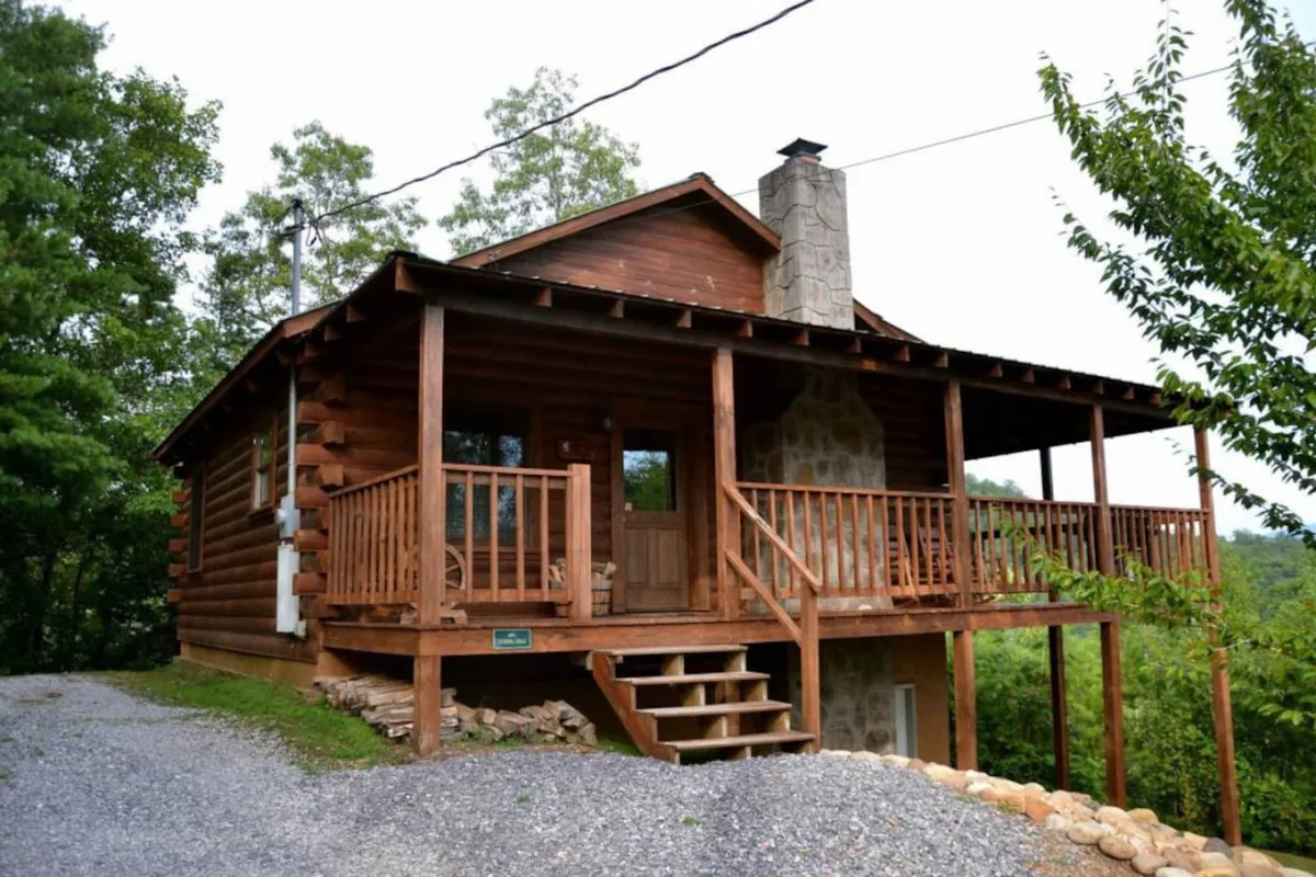 A cozy log cabin exterior with a stone chimney, inviting front porch, and lush greenery surrounding the property. The wooden porch suggests a welcoming space for relaxation, with ample natural light and scenic views.