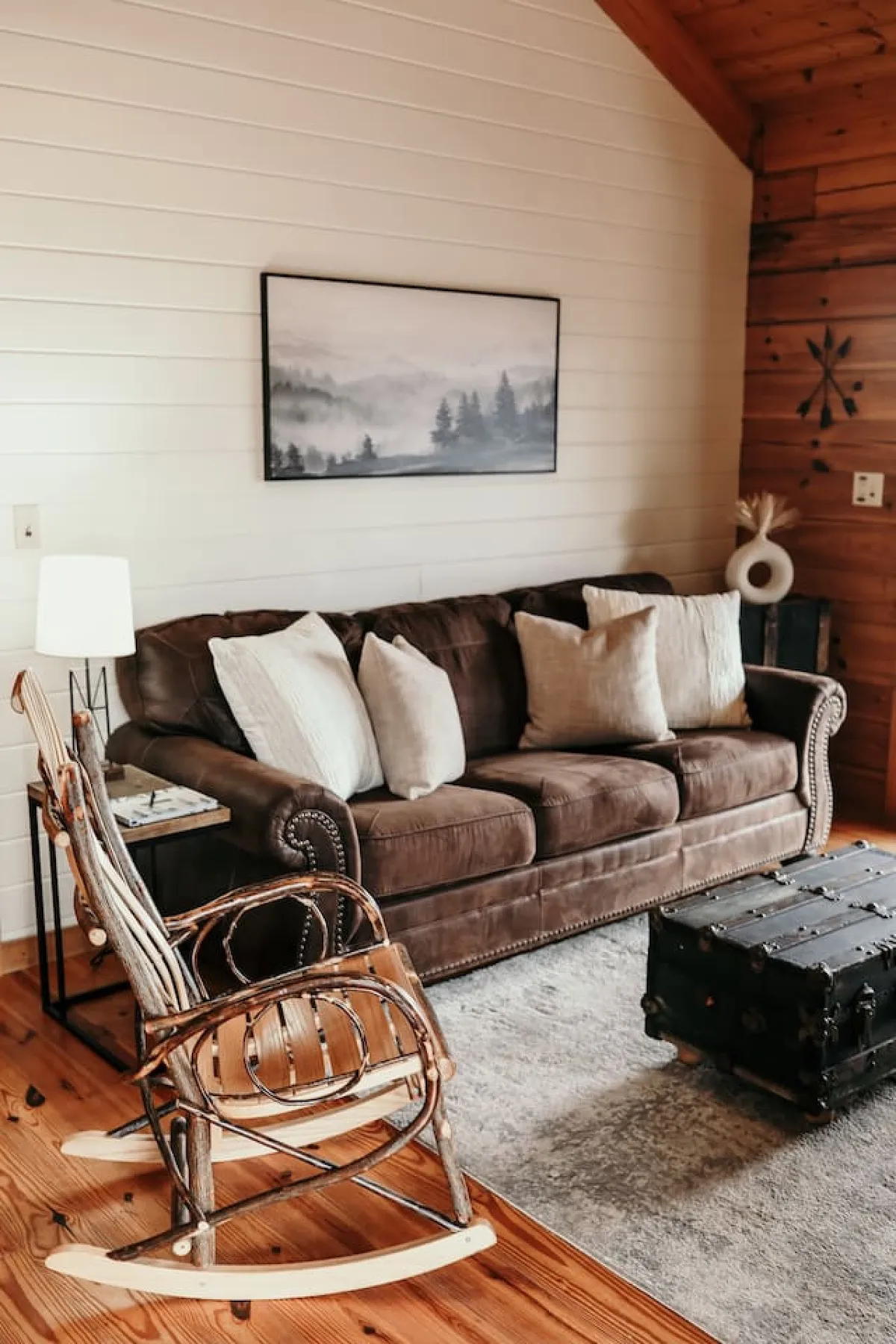 A secondary interior space with a comfortable brown sofa, a rocking chair, and a black trunk, accented by a landscape painting on the wall. The mood is inviting and serene, ideal for relaxing, reading, or quiet conversation in a den-like atmosphere.