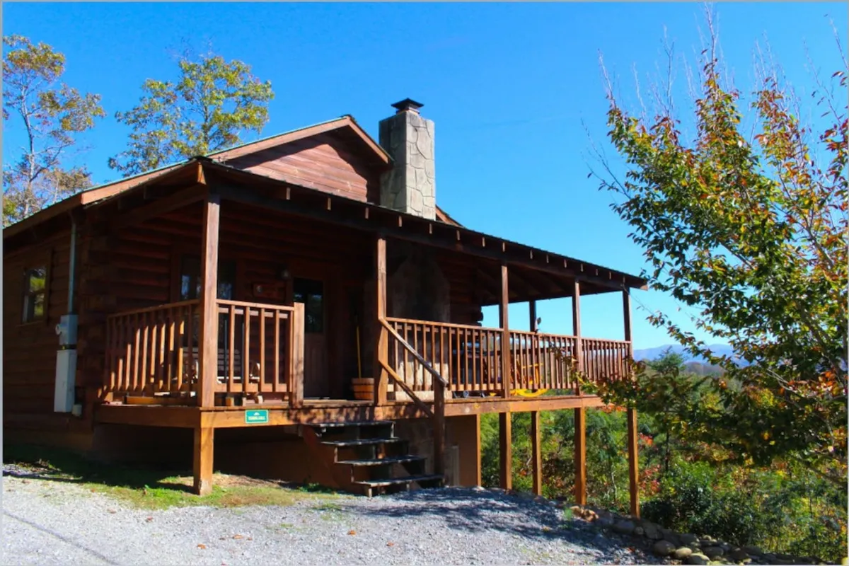 The image showcases a charming log cabin perched among whispering trees, with a welcoming front porch perfect for morning coffee and a stately stone chimney hinting at cozy nights by the fire. Sunlight dapples the wood exterior, inviting guests to relax and savor the peaceful mountain setting.
