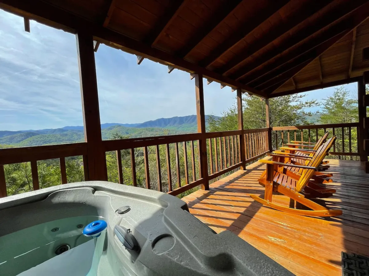 A warm, inviting exterior shot showcases a bubbling hot tub and wooden chairs with a sweeping mountain view beyond. The scene is tranquil, perfect for unwinding after a day of exploration, with ample space for socializing or a quiet moment with nature.