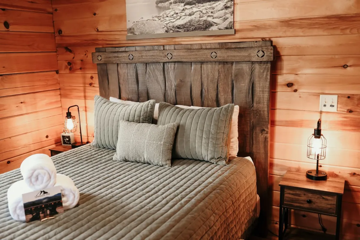 A cozy bedroom space with a wooden headboard, a bed made up with a textured quilt and decorative pillows, and lantern-style bedside lamps. The natural light filtering in creates a warm, restful vibe.