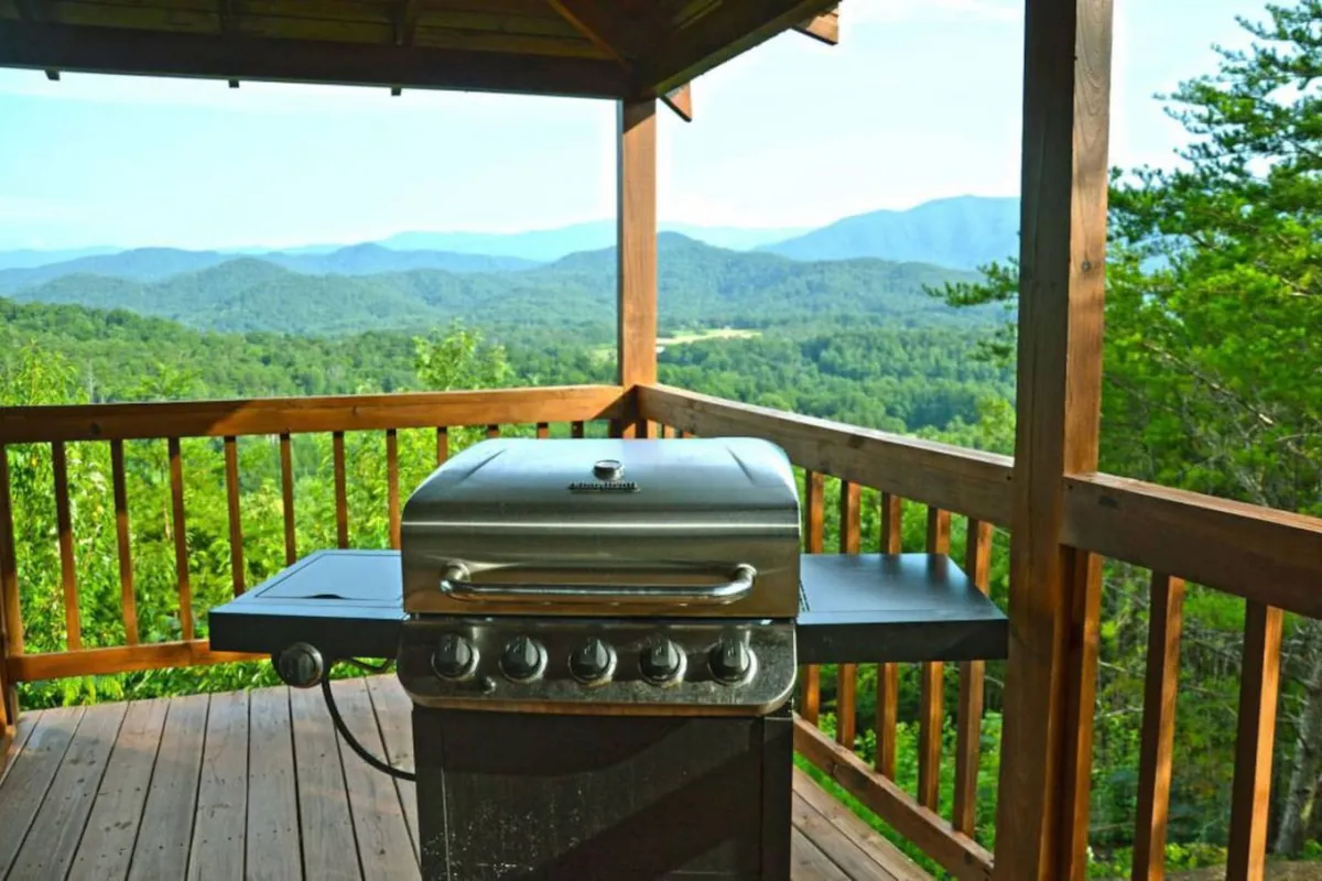 An outdoor deck scene with a grill in the foreground, mountain views stretching out beyond the railing. The deck is set up for social gatherings, with ample space for barbecuing and enjoying the scenery. The setting sun casts a warm glow, enhancing the relaxed, communal vibe.