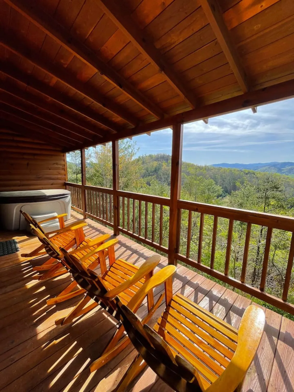 The scene captures a tranquil deck setting with two rocking chairs and a hot tub positioned to provide uninterrupted views of the surrounding mountains. The morning or evening light bathes the space in a soft, inviting glow, reinforcing a sense of peace and seclusion.