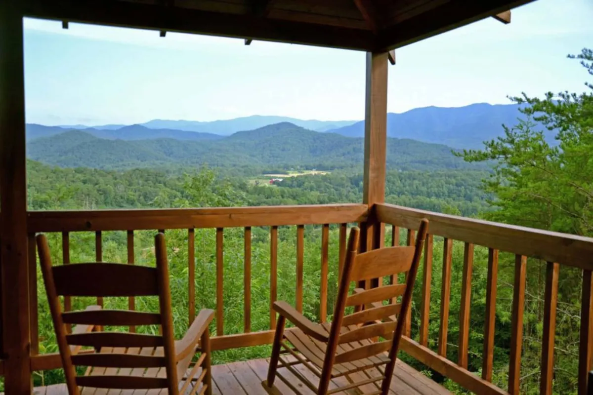 A pair of wooden rocking chairs sit on a spacious deck overlooking a sweeping mountain vista, framed by a rustic wooden railing. The early morning light casts a warm glow over the scene, inviting relaxation and quiet contemplation. Forested slopes roll away into the distance, creating a sense of seclusion and connection to nature.