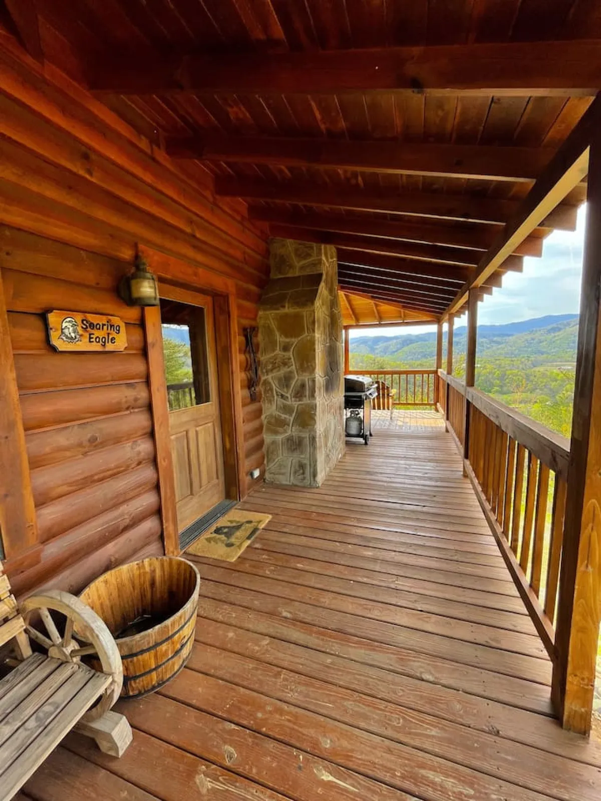 This image features a welcoming wooden porch with a stone fireplace, a cozy bench, and a classic barrel, all set against a backdrop of beautiful mountain views. The space suggests a perfect spot for families and pets to relax after a day of adventure, with ample outdoor seating and a sense of connection to nature.