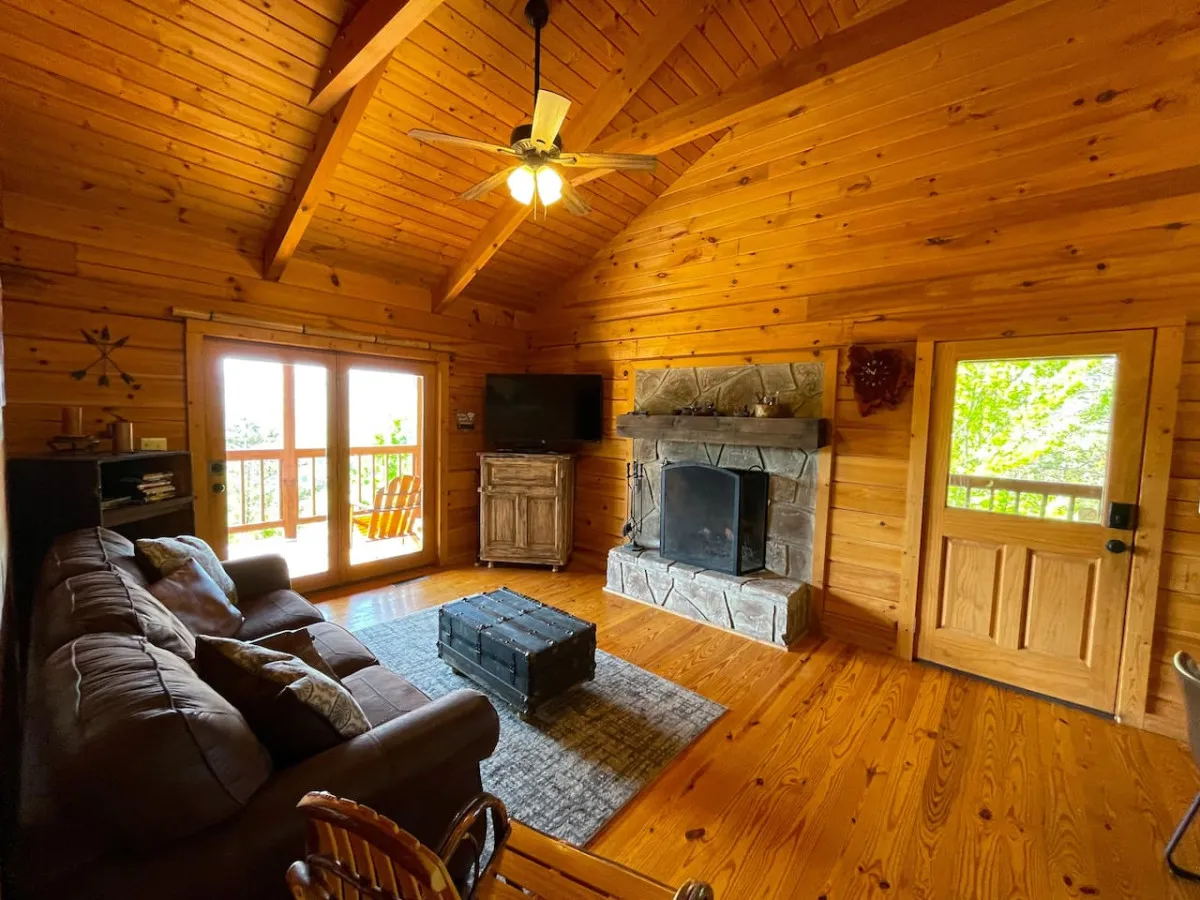 The image showcases a cozy living room with a real-wood fireplace at its center, plush sofas inviting you to relax, a wooden door adding rustic charm, and large windows that reveal stunning views of the Smoky Mountains. The natural light streaming in highlights the comfortable, open layout ideal for gatherings or quiet evenings, with a smart TV visible for entertainment.