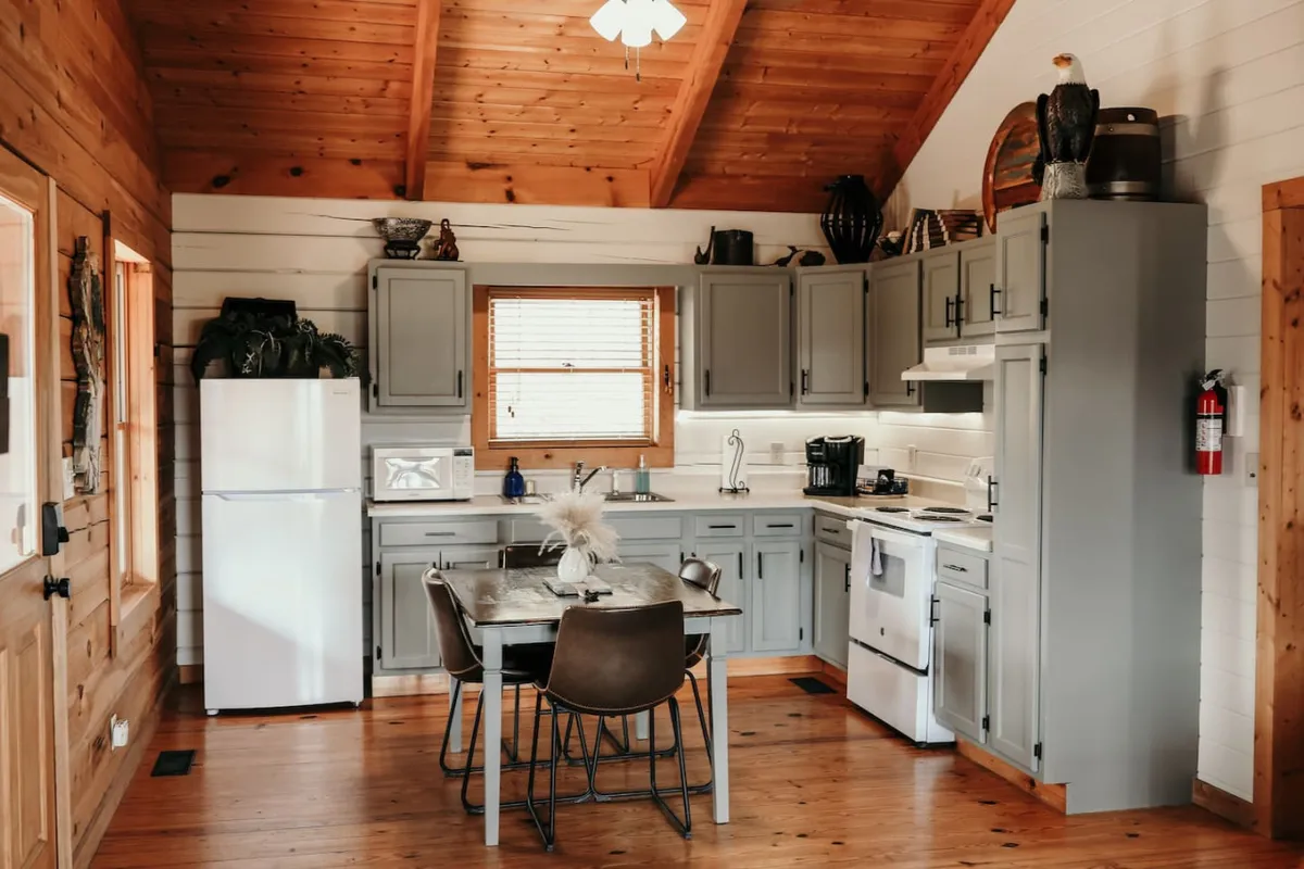 A warm, welcoming kitchen interior featuring a modern kitchen table set close to a large window. The full-size refrigerator and oven are visible, with the table ideally positioned to overlook the scenic outdoors. The natural light pouring through the windows enhances the inviting atmosphere, perfect for family meals and gatherings.