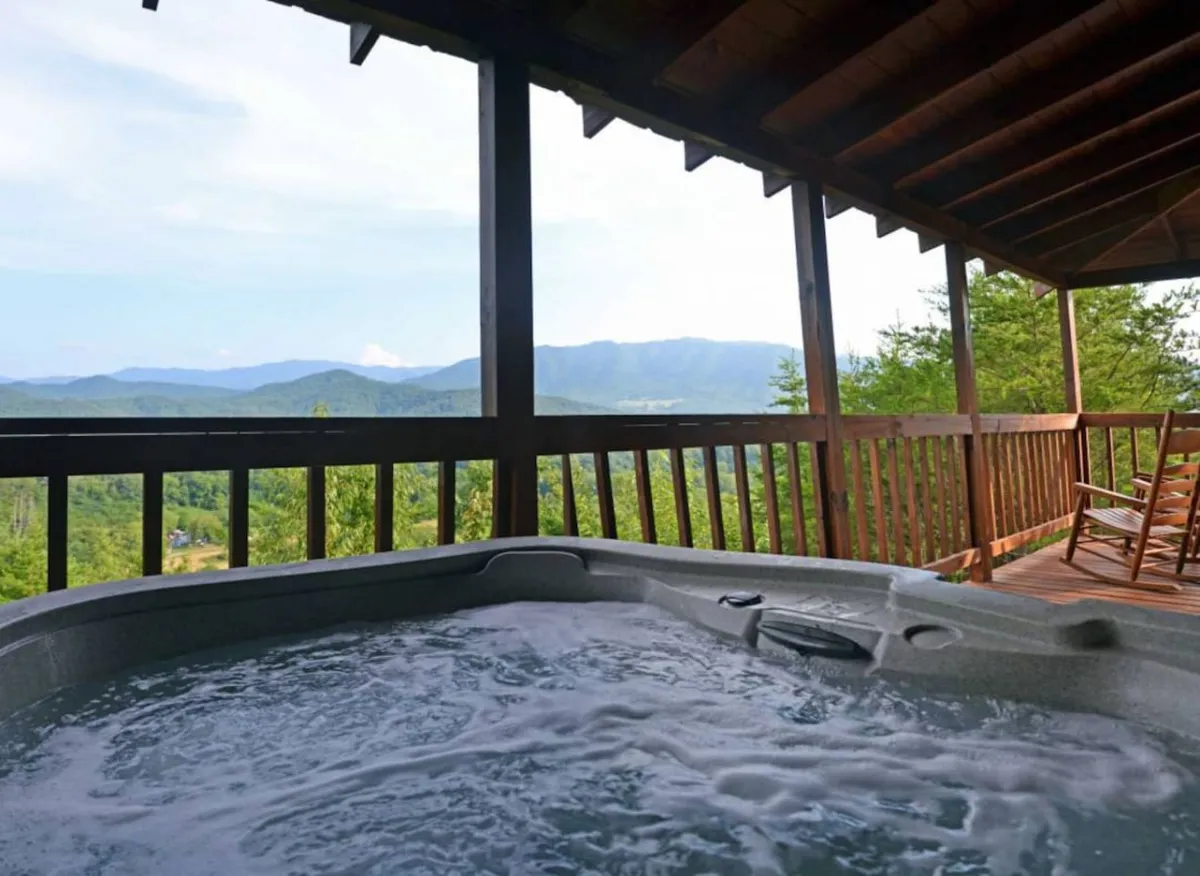 This image features a spacious wooden deck with a bubbling hot tub overlooking a sweeping mountain view. The foreground includes a sturdy wooden railing and cozy rocking chairs, inviting guests to relax and enjoy the tranquil atmosphere. The setting sun casts a warm glow over the landscape, enhancing the sense of retreat and romance.