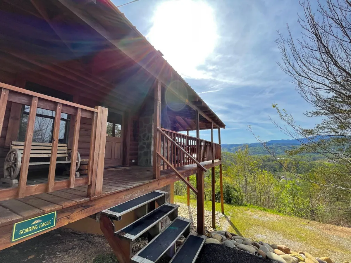 A sunlit wooden porch features a classic rocking chair facing out toward the misty silhouette of mountains in the distance. Gentle sunlight flares through the background, suggesting the tranquility of morning or evening. The porch’s vantage point highlights the connection between the cabin’s comfortable spaces and the natural surroundings.